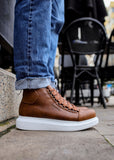 A person wearing brown high-top sneakers with white soles and laced up, standing on a stone surface with black chairs and a table in the background.