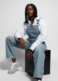 Person wearing a denim overalls and grey Apollo Moda sneakers sitting on a black cube against a light gray background